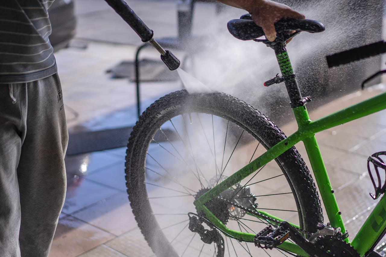 A man cleans a green bicycle with a hose, focusing on the wheel and frame outdoors.
